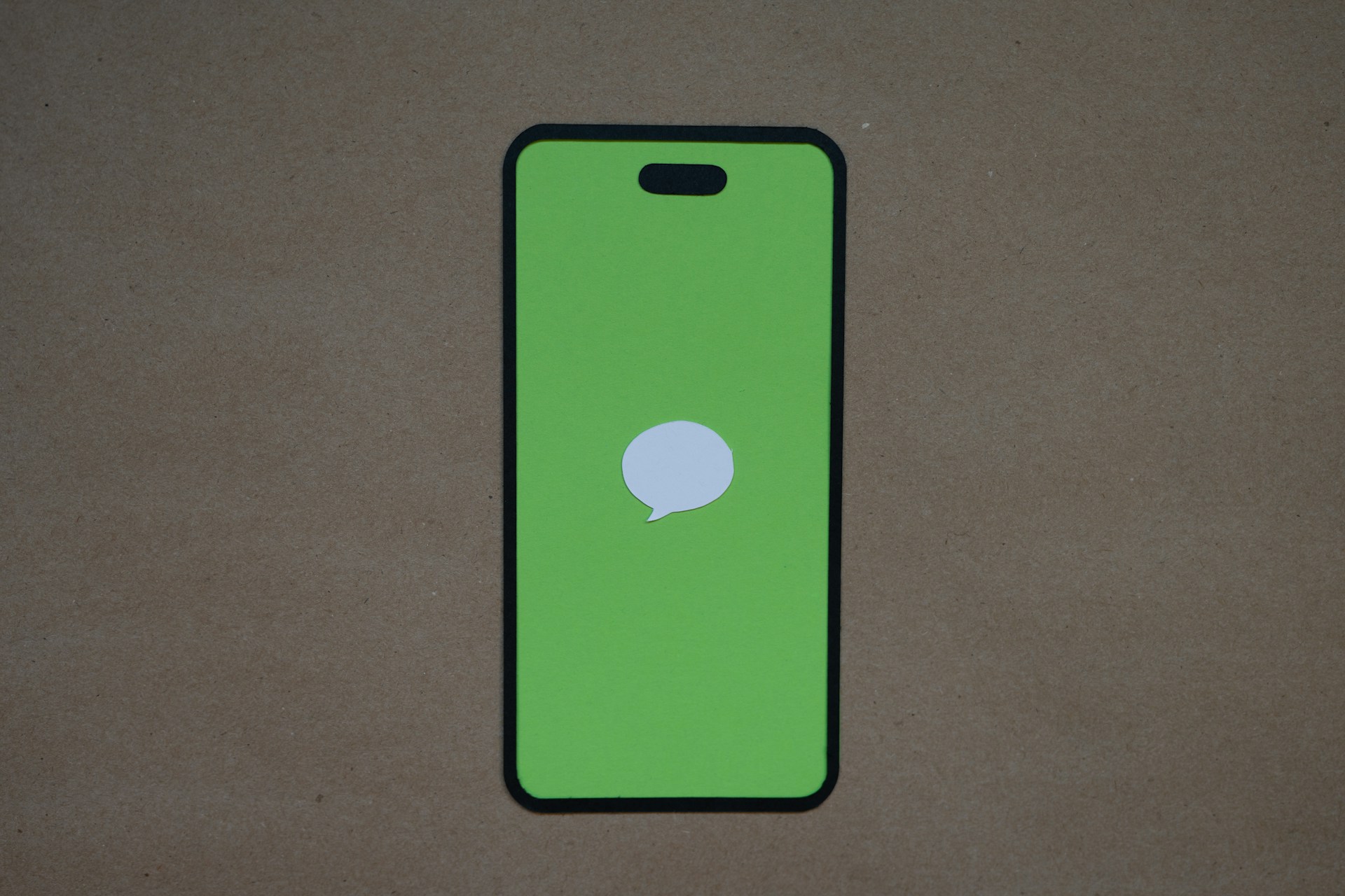 Smartphone silhouette with a bright green screen and a centered white speech-bubble icon suggesting a messaging notification, placed on a brown kraft-paper background.