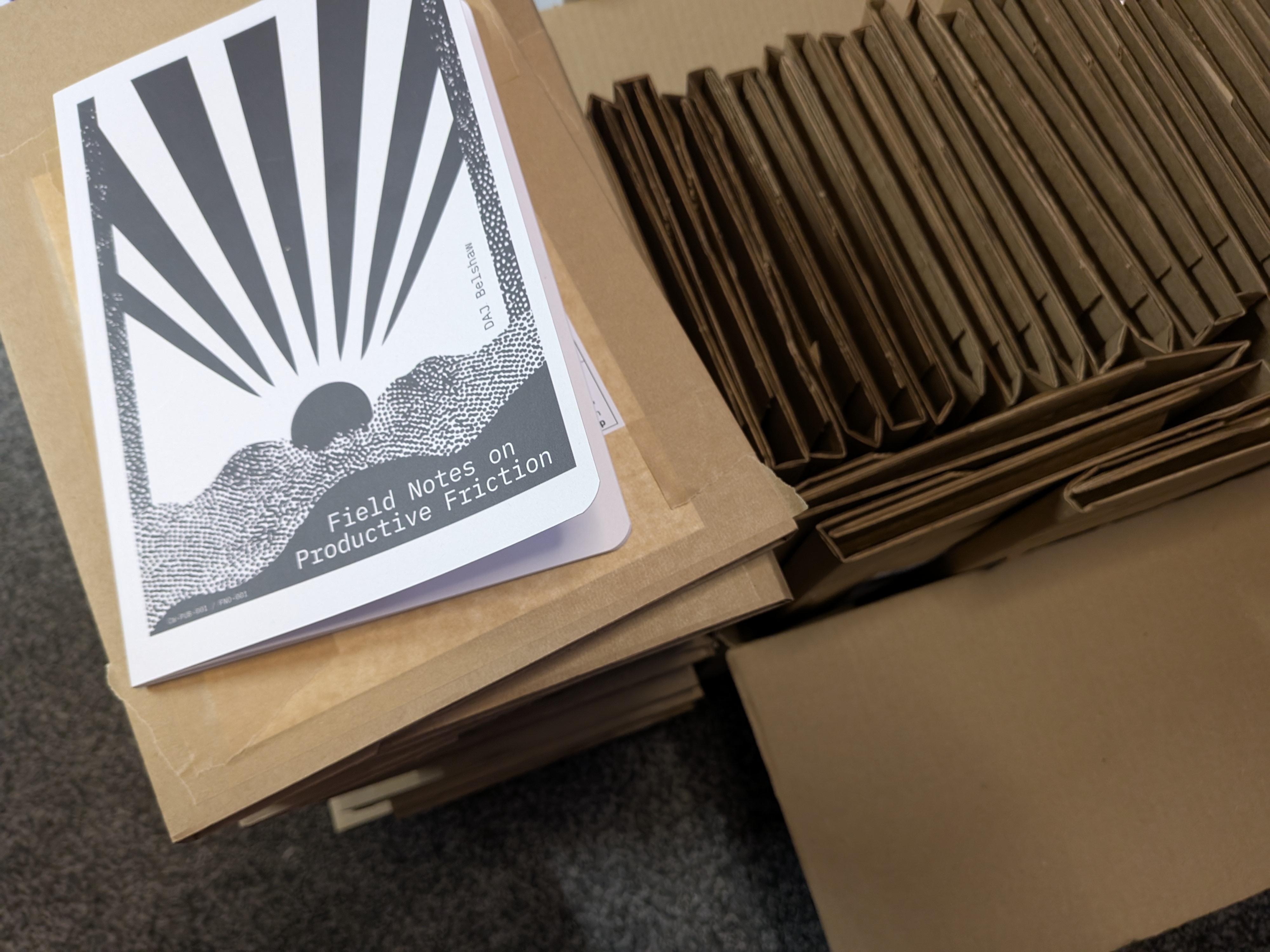 Stack of zines titled Field Notes on Productive Friction with a sunburst cover illustration, resting on flattened cardboard and a neatly stacked pile of folded brown shipping envelopes ready for mailing.