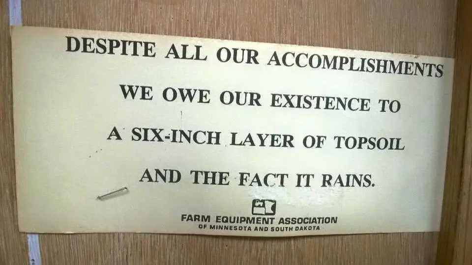 Humorous framed sign from the Farm Equipment Association of Minnesota and South Dakota reading: 'Despite all our accomplishments we owe our existence to a six-inch layer of topsoil and the fact it rains.'