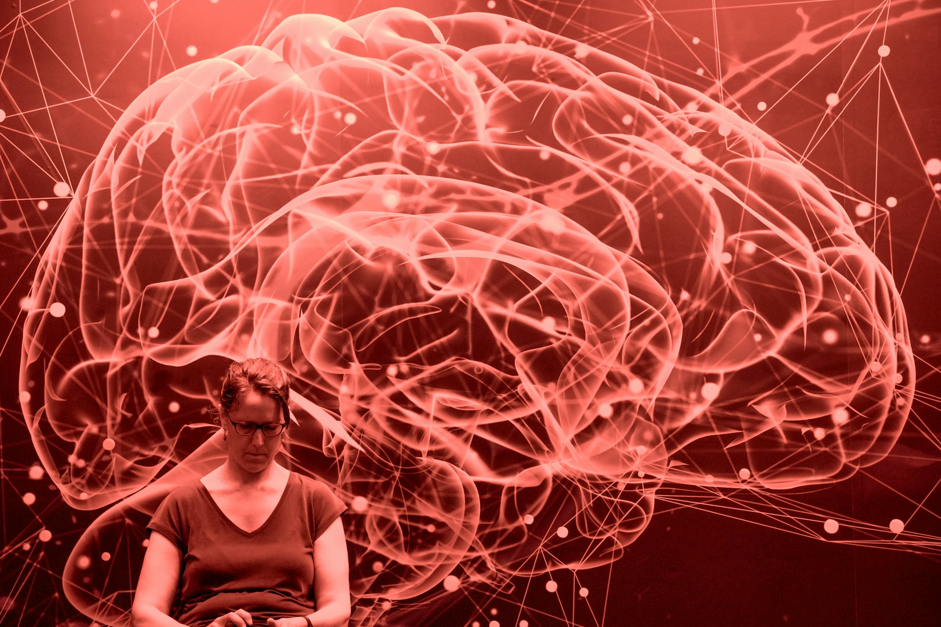 Red tinted photograph of a large stylised brain wall art with a woman using her phone in front of it.
