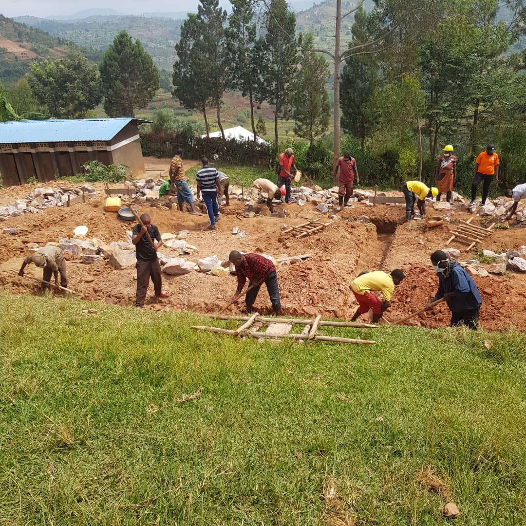 Community workers digging and leveling a large foundation trench, shoveling soil and moving rocks on a rural hillside site with trees and a blue-roofed building in the background.