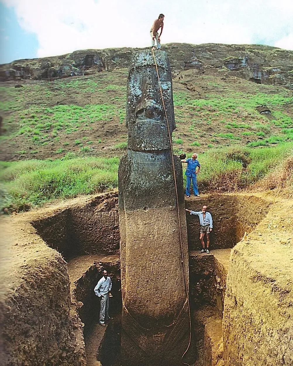 Partially excavated Easter Island moai statue standing upright in a deep pit, with archaeologists and ropes revealing its buried body and showing scale.
