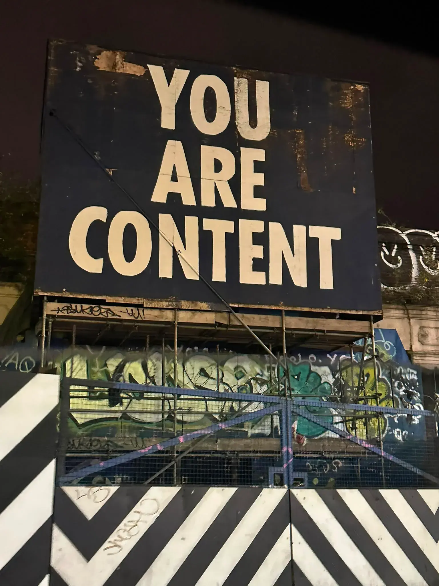 Bold street-art billboard reading 'YOU ARE CONTENT' in large white letters on a weathered black panel above graffiti-covered scaffolding and black-and-white chevron barriers at night.