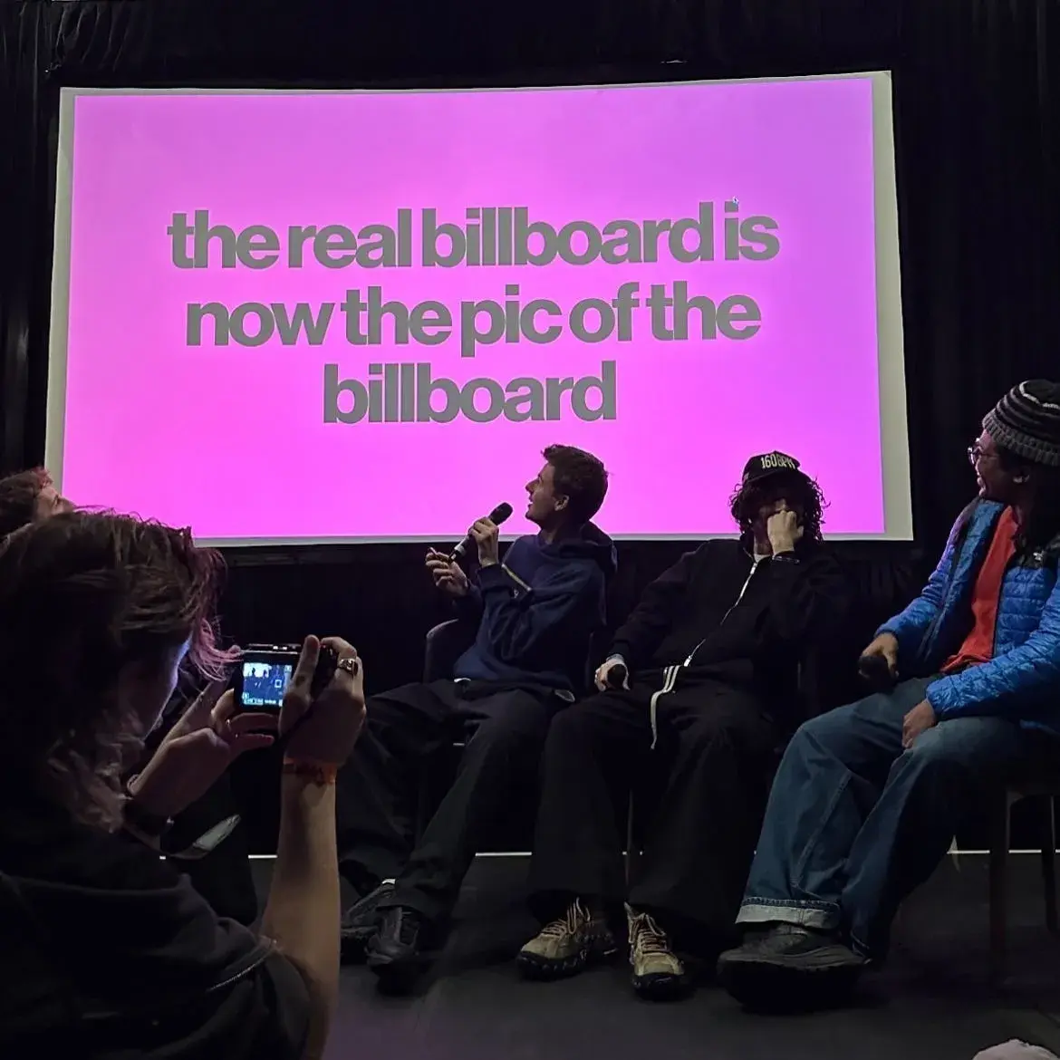 Someone photographing people in a panel session. One holds a microphone and looks up at a screenwhich reads "the real billboard is now the pic of the billboard"